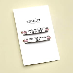 May include: A white card with the word "amulet" and the text "personalised shoe tags". Two silver shoe tags are displayed, one with "I DON'T KNOW ABOUT YOU" and the other with "BUT I'M FEELING 26.2". Both tags have pink cords.