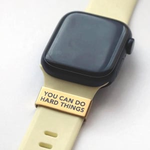 Peut inclure: Une montre intelligente noire avec un bracelet crème. Un connecteur de bracelet en métal doré avec le texte "YOU CAN DO HARD THINGS" est fixé au bracelet. Le cadran de la montre est noir.