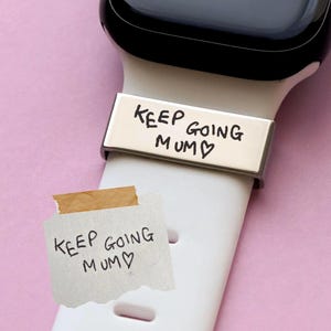 Può includere: Cinturino d'orologio argentato con la scritta "KEEP GOING MUM". Un cinturino bianco è attaccato a uno smartwatch nero. Un piccolo pezzo di carta con lo stesso testo è attaccato al cinturino. Lo sfondo è rosa.