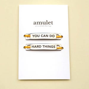 Peut inclure: Une carte blanche présente deux paires d'étiquettes de chaussures argentées avec des lacets jaunes. L'étiquette supérieure indique "YOU CAN DO" et l'étiquette inférieure indique "HARD THINGS". Le mot "amulet" et "personalised shoe tags" sont imprimés en haut de la carte.