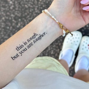 Puede incluir: Primer plano de un antebrazo con un tatuaje temporal que dice: "this is tough, but you are tougher". Una pulsera de oro y perlas se lleva en la muñeca. Se ven zuecos blancos y una falda amarilla.