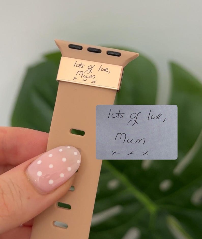 May include: A tan Apple Watch band with a gold-colored metal accent. The metal accent has the handwritten text "lots of love, Mum" engraved on it. A small gray sticky note with the same text is also visible.