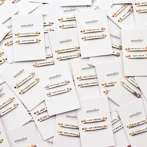 May include: A collection of white rectangular display cards, each branded with the word amulet, featuring two small metallic pins with engraved motivational phrases such as You've Got This, Keep Going, I Am Strong, and Strava Kudos.