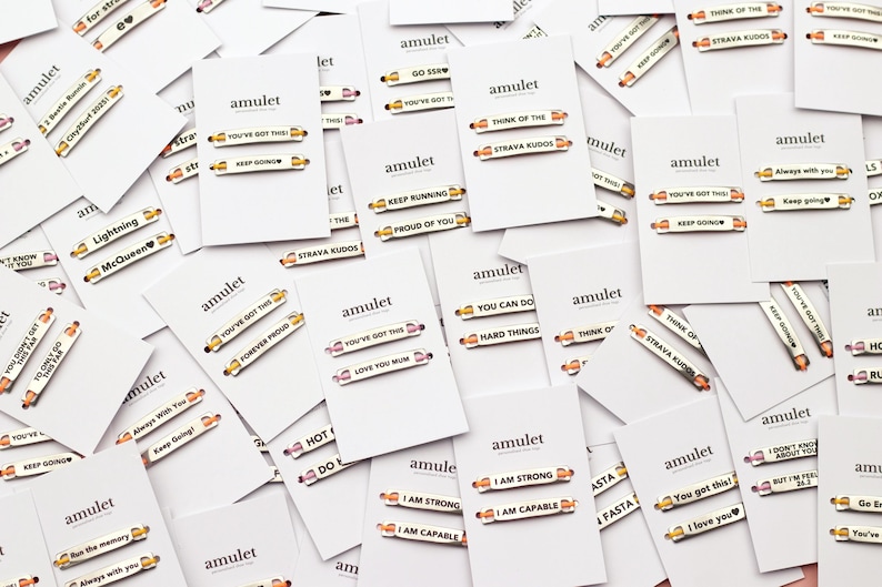 May include: A collection of white rectangular display cards, each branded with the word amulet, featuring two small metallic pins with engraved motivational phrases such as You've Got This, Keep Going, I Am Strong, and Strava Kudos.