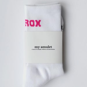 May include: White socks with the word "ROX" in pink letters at the top. The socks are folded and wrapped with a white band that reads "my amulet" and "a beacon of strength, resilience and determination".