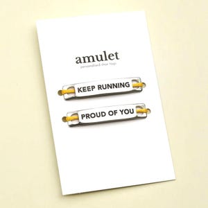 Op de afbeelding: Een witte kaart met het woord "amulet" en de tekst "personalised shoe tags" in het zwart gedrukt. Er worden twee zilveren schoenlabels met gele veters weergegeven. Eén label leest "KEEP RUNNING" en de andere leest "PROUD OF YOU".