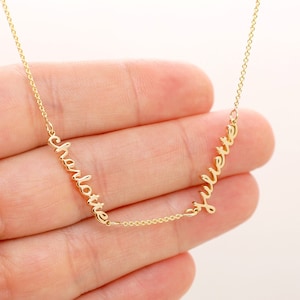 May include: A delicate gold necklace with the names "Charlotte" and "Juliette" in cursive script. The names are linked by a fine chain, creating a personalised jewellery piece. The necklace is shown against a soft background.