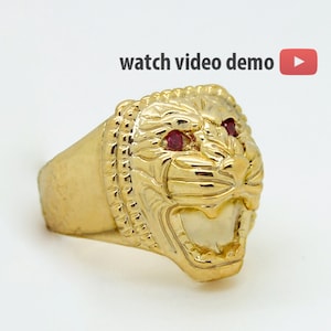 Könnte beinhalten: Goldener Löwenkopfring mit roten Edelsteinaugen. Der Ring ist mit einem strukturierten Design und einem brüllenden Löwenkopf verziert.