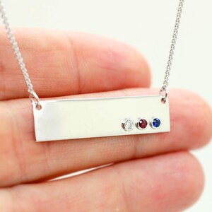 A hand holding a silver necklace with a rectangular pendant featuring three gemstones.