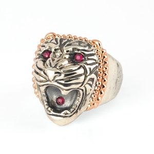 May include: A silver ring with a detailed tiger head design. The tiger's eyes are set with red gemstones and the ring is accented with a rose gold beaded band.