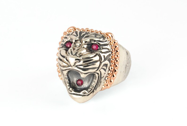 May include: A silver ring with a detailed tiger head design. The tiger's eyes are set with red gemstones and the ring is accented with a rose gold beaded band.