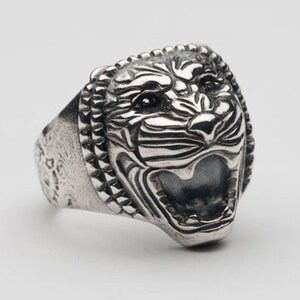 May include: A silver ring with a detailed tiger head design. The tiger's mouth is open and its teeth are visible. The ring is made of metal and has a textured surface.