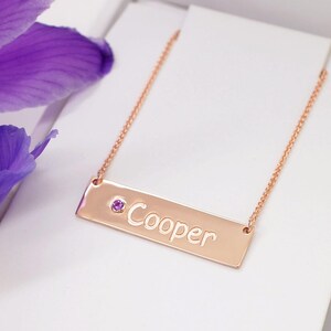 A gold-colored necklace with a rectangular pendant engraved with the name "Cooper" and a small purple gemstone.