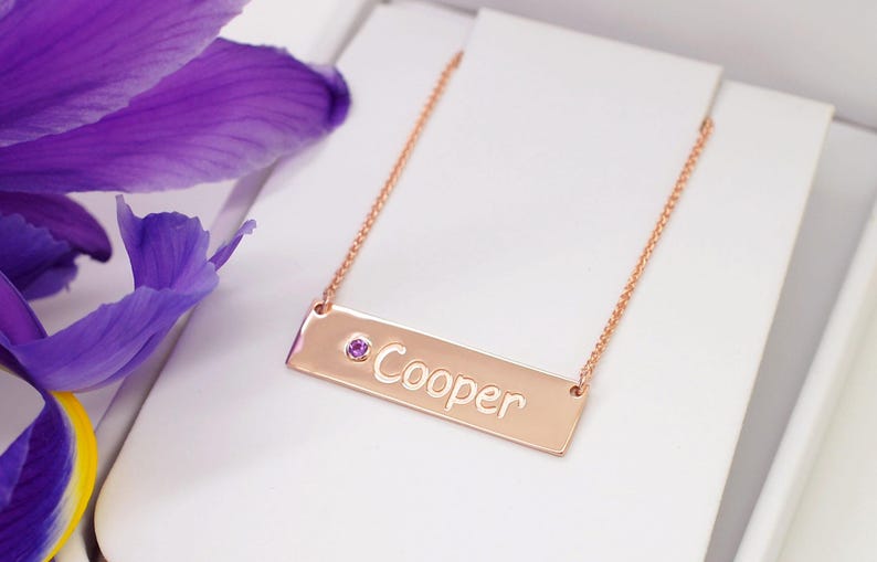 A gold-colored necklace with a rectangular pendant engraved with the name "Cooper" and a small purple gemstone.