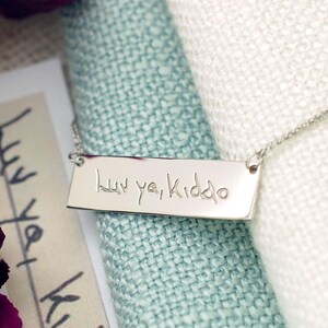A silver necklace with a rectangular pendant engraved with the text "luu yi Kido" is displayed on a white fabric surface.
