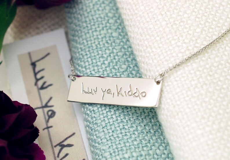 A silver necklace with a rectangular pendant engraved with the text "luu yi Kido" is displayed on a white fabric surface.