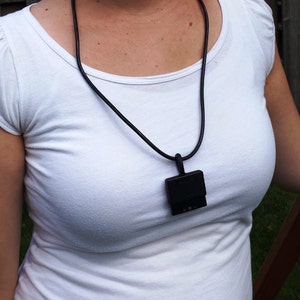 May include: A black lanyard with a square-shaped pendant. The pendant has a textured surface and a small opening on the top.