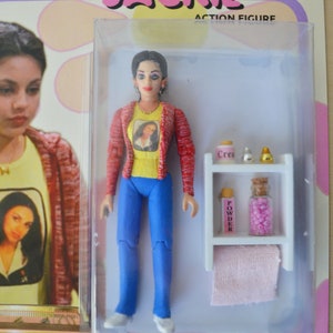 May include: A plastic action figure of a young woman with brown hair and a red and white striped shirt. The figure is holding a small bottle of perfume. The figure is packaged in a clear plastic box with a white background. The box has the words "Jackie Action Figure" printed on it.