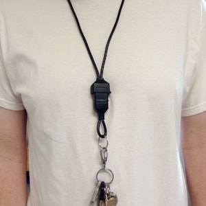 May include: A black lanyard with a black plastic clip and a silver key ring with multiple keys attached. The lanyard is worn around the neck of a person wearing a white t-shirt.