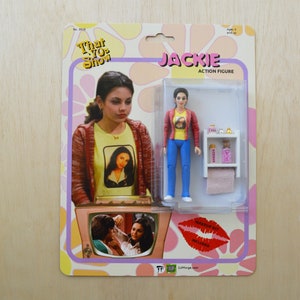 May include: A retro-style action figure of Jackie from the TV show "That 70s Show". The figure is wearing a red shirt, blue jeans, and a yellow shirt with a picture of Jackie on it. The figure is holding a small bottle of perfume and a towel. The figure is packaged in a cardboard box with a picture of Jackie on the front. The box also includes the text "Jackie Action Figure" and "Makeup Kit Included".