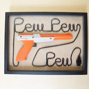 May include: A framed piece of art featuring a black wire shaped like the word "Pew" with a NES Zapper gun in the middle. The background is a brown burlap fabric.