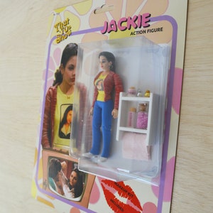 May include: A plastic action figure of Jackie from the TV show That 70s Show. The figure is wearing a red sweater, blue jeans, and a yellow t-shirt with a picture of a woman on it. The figure is packaged in a clear plastic blister pack with a white background and a pink and yellow floral design. The packaging also includes a makeup kit.