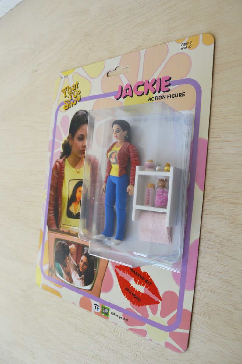 May include: A plastic action figure of Jackie from the TV show That 70s Show. The figure is wearing a red sweater, blue jeans, and a yellow t-shirt with a picture of a woman on it. The figure is packaged in a clear plastic blister pack with a white background and a pink and yellow floral design. The packaging also includes a makeup kit.