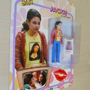 May include: A retro-style action figure of Jackie from the TV show "That '70s Show". The figure is wearing a red jacket, yellow shirt, and blue jeans. The packaging includes a small plastic stand and a makeup kit.