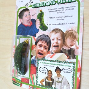 以下が含まれることがあります： クリスマスのピクルスのオーナメント、緑色のキラキラしたピクルスが、子供がおもちゃのピクルスを持っている写真と「Hey Kids, It's the Christmas Pickle」という文字が書かれた白いカードに飾られています。カードには、泣いている3人の子供の写真と「A Time Honored German Tradition」という文字も書かれています。