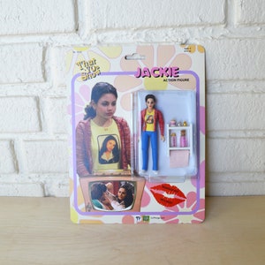 May include: A retro-style action figure of Jackie from the TV show "That '70s Show". The figure is wearing a red shirt, blue jeans, and a yellow shirt with a picture of Mona Lisa on it. The figure comes with a small makeup set and a pink towel.