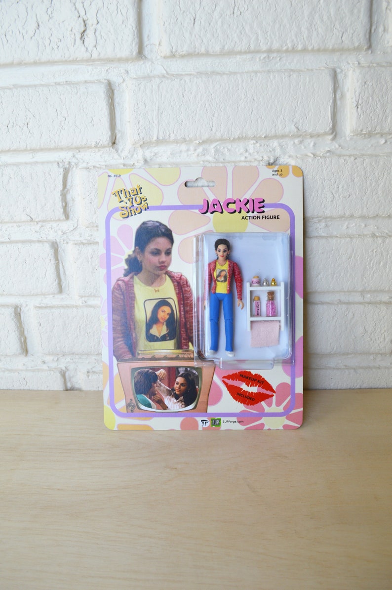 May include: A retro-style action figure of Jackie from the TV show "That '70s Show". The figure is wearing a red shirt, blue jeans, and a yellow shirt with a picture of Mona Lisa on it. The figure comes with a small makeup set and a pink towel.