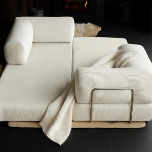 May include: Cream-colored sectional sofa with a corduroy texture. The sofa features a rounded armrest and backrest cushions. A cream-colored throw blanket is draped over the sofa. The sofa sits on a light brown rug.
