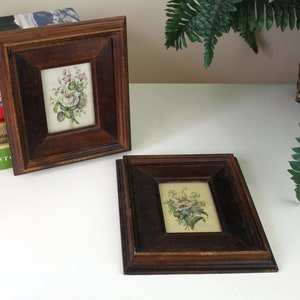 May include: Two small, dark wood framed pictures with floral prints. The pictures are on a white surface with a green plant in the background.
