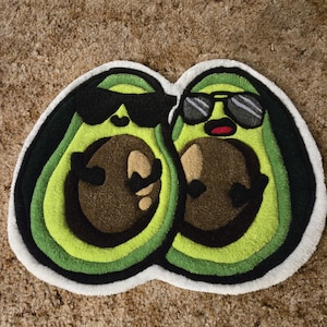 May include: A fun avocado-shaped rug depicting two smiling avocado halves wearing sunglasses. The rug has a white border, with green, black, and brown details. The rug is designed to look like a pair of avocados.