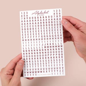 Könnte beinhalten: Ein Bogen mit burgunderfarbenen Alphabet-Aufklebern, der Buchstaben, Zahlen und Symbole enthält. Das Wort "Alphabet" ist kursiv geschrieben. Die Aufkleber werden von Händen gehalten.