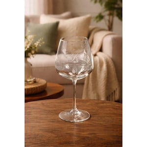 May include: A clear wine glass with etched floral designs. The glass has a long stem and a wide bowl, sitting on a dark wooden table. The background includes a sofa with pillows and a blanket, suggesting a cozy interior setting.