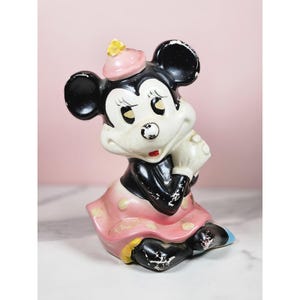 Może przedstawiać: Figurka Minnie Mouse w stylu vintage w pozycji siedzącej. Ceramiczna figurka ma czarne uszy, czarną sukienkę i różową spódnicę w kropki. Nosi różowy kapelusz z żółtym kwiatem i białe rękawiczki. Figurka nosi ślady użytkowania.