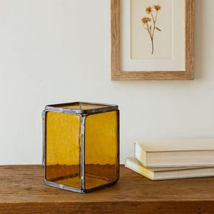 May include: A square, amber-colored glass lantern with a metal frame. The lantern is sitting on a wooden surface, with a framed botanical print and a stack of books in the background. The lantern is a decorative home accent.