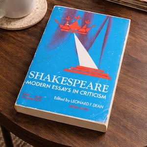 Puede incluir: Un libro de bolsillo titulado Shakespeare: Modern Essays in Criticism, editado por Leonard F. Dean. La portada es de color azul brillante con un gráfico rojo de una corona sobre un triángulo blanco y escalones rojos. El texto está en blanco y rojo.