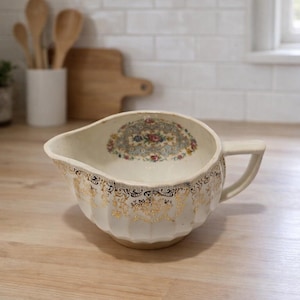 May include: Cream-colored gravy boat with a floral design inside and gold filigree around the rim. The boat has a handle and a spout for easy pouring. A vintage kitchenware item.