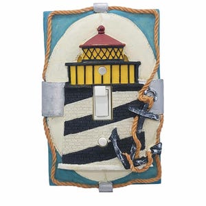 May include: A rectangular light switch cover plate featuring a raised lighthouse design with black and white stripes, a yellow lantern room, and a red roof. A black anchor wrapped in a tan rope is attached to the side, set against a blue background.