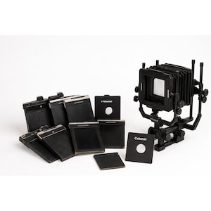 May include: A black large format view camera with a bellows and a grid focusing screen, accompanied by several black film holders and lens boards. The components feature the brand name Calumet printed in white text on the black surfaces.
