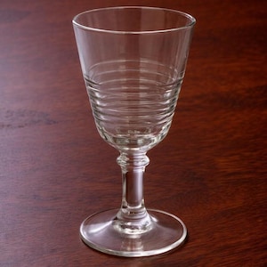 May include: A clear wine glass with a wide bowl and a ridged pattern on the lower section. It has a round base and a slender stem. The glass is empty and is placed on a dark wooden surface. The glass is suitable for wine.