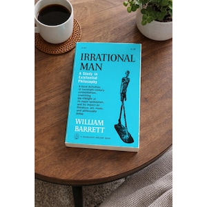 Może przedstawiać: Książka "Irrational Man" Williama Barretta, z jasnoniebieską okładką i grafiką przedstawiającą osobę. Książka leży na drewnianym stole obok filiżanki kawy i małej rośliny.