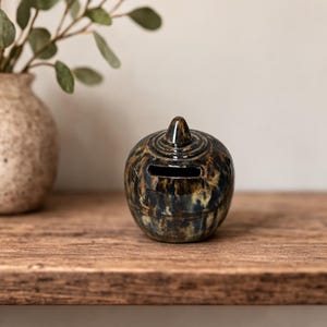 Pode incluir: Um cofrinho de cerâmica com uma cobertura escura e manchada em tons de castanho, azul e dourado. O banco redondo tem uma ranhura para moedas e um pequeno topo pontiagudo. Está sobre uma superfície de madeira, com um vaso e vegetação no fundo.