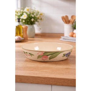 以下が含まれることがあります： ピンクのチューリップと緑の葉のフローラルデザインが施された、クリーム色の丸いセラミック製のベーキングディッシュ。木製の表面に置かれ、背景にはぼやけたキッチン用品があります。