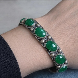 May include: A silver-toned cuff bracelet featuring a series of oval-shaped, smooth green stones set in a row. The metal band displays a twisted rope texture along the edges and small spherical metal beads positioned between each green stone.