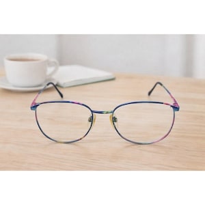 May include: Round-framed eyeglasses with a blue metal frame and colorful floral accents on the temples. The glasses are set on a light wood surface, with a white coffee cup and a notepad in the background.
