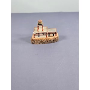 Può includere: Una statuetta di un faro in miniatura con una torre rossa e bianca e una base rettangolare. Il faro è posto su una base di legno con una superficie marrone strutturata. L'edificio ha un esterno bianco con finiture rosse.