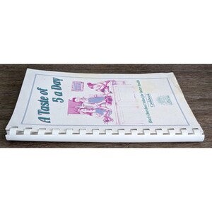 May include: A spiral-bound cookbook titled "A Taste of 5 a Day" with a vintage illustration on the cover. The cover features text in blue and a black and white illustration of people. The cookbook is open on a wooden surface.